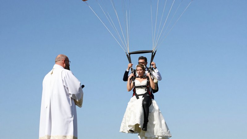 Bride in wedding dress paragliding with groom approaches a waiting wedding officiant under a clea...