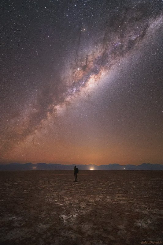 Silhouetted hiker stands on a dry desert salt flat gazing up at the brilliant arching Milky Way a...