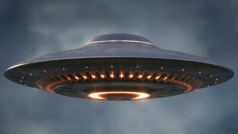 Classic metallic flying saucer UFO with glowing orange lights hovers against a moody overcast gray sky