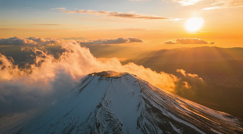 Aerial view of snow-capped Mount Fuji rising above glowing golden clouds at warm sunrise, bright sun low on the orange hor...
