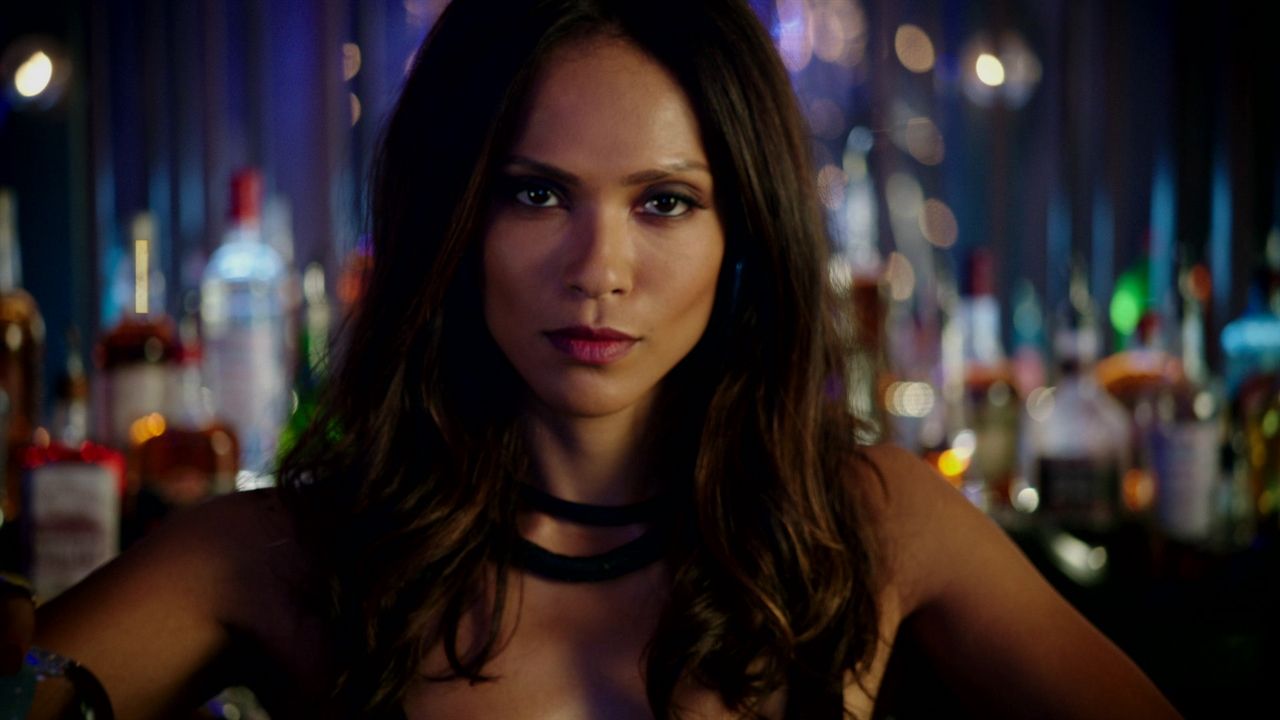 Woman bartender in a dimly lit bar with bottles and bokeh lights in the background.
