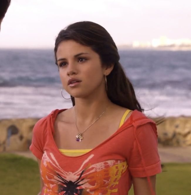Young woman by the sea in an orange graphic tee with yellow straps and hoop earrings.