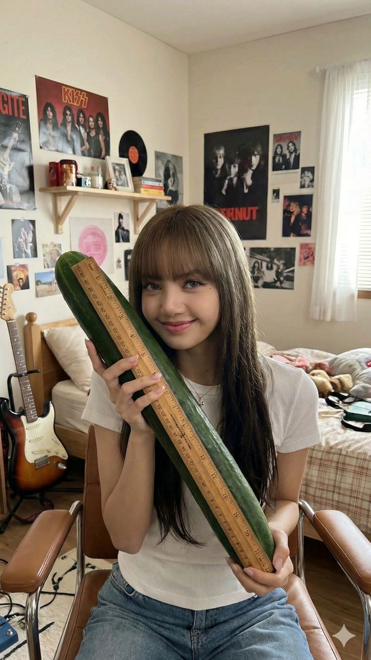 Smiling girl holding a large cucumber with a ruler in a guitar-filled bedroom.