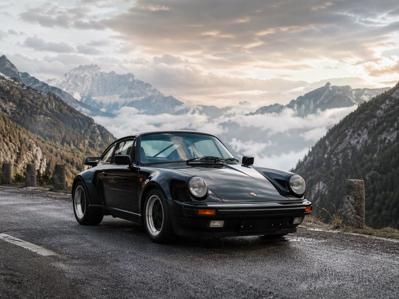 Black vintage Porsche 911 parked on a mountain road with misty valley and alpine mountains in the...
