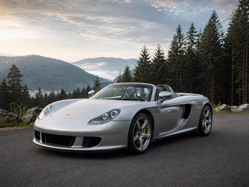Silver Porsche convertible parked on a mountain road with pine forest background.