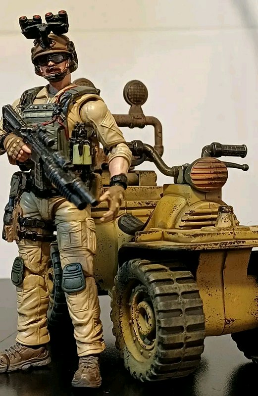 Desert-themed military action figure with night-vision goggles beside a weathered yellow quad bike.