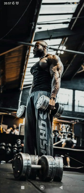 Muscular man in a sleeveless shirt lifting heavy dumbbells in a gritty indoor gym.