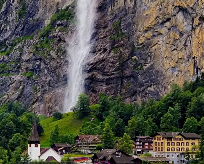 Alpine village with a tall waterfall cascading down a rocky cliff behind green trees.