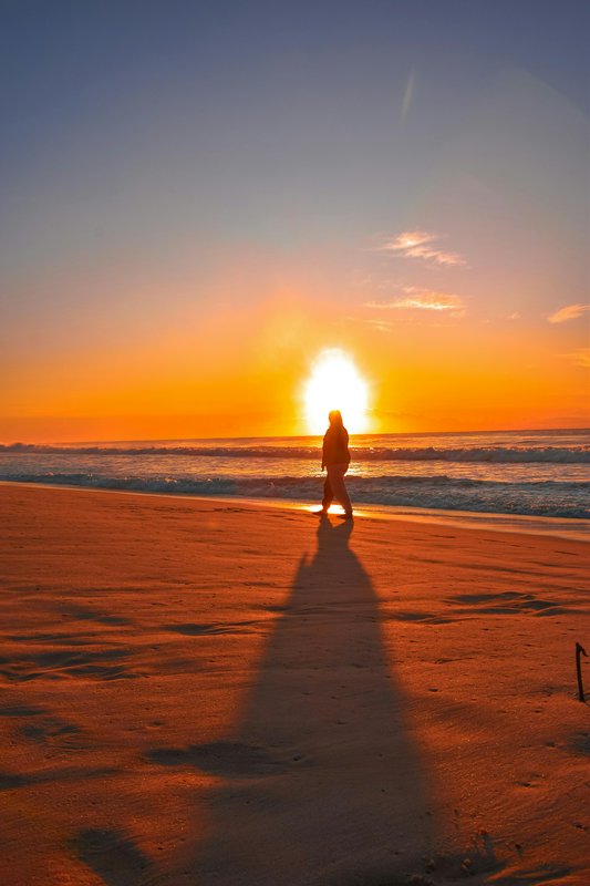 Silhouette of a person walking on a sandy beach at sunset with a long shadow.