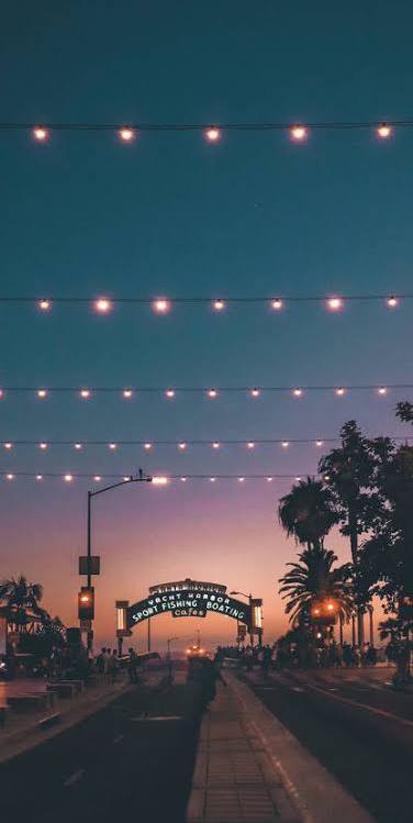 Evening street scene with string lights overhead, palm trees, and a glowing archway sign at sunset.