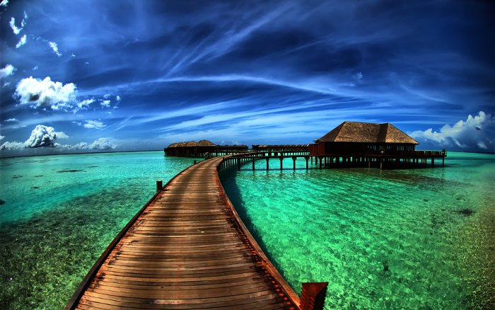 Wooden boardwalk leading to overwater bungalow villas in a turquoise lagoon under a vivid blue sky.