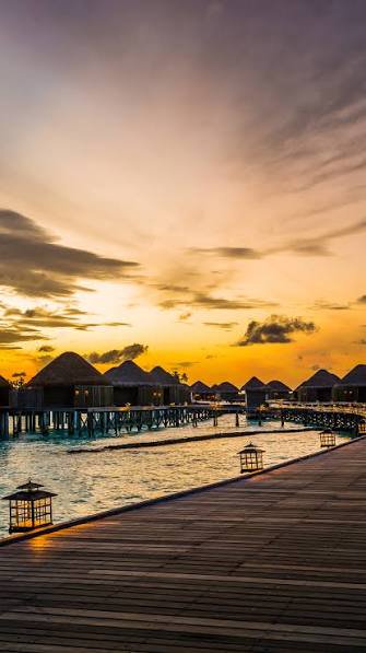 Golden sunset sky over luxury overwater bungalow resort, wooden boardwalk lined with glowing lant...