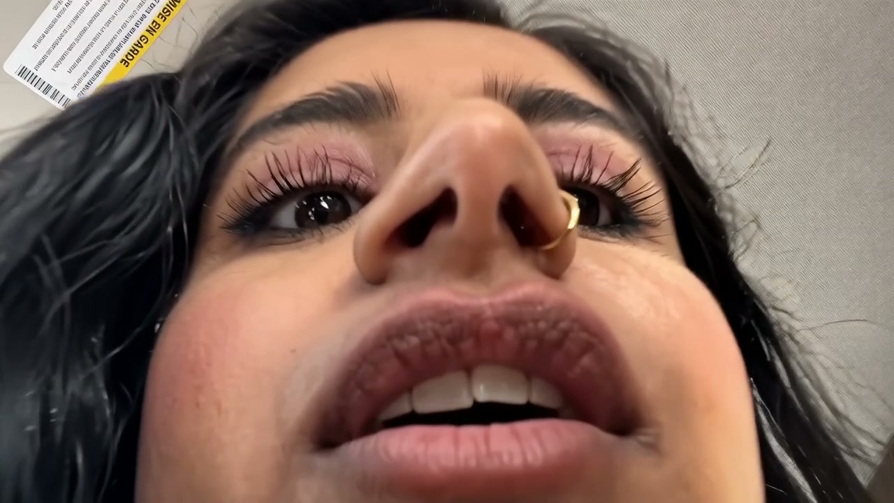 Close-up of a woman's face from below, featuring a gold nose ring, pink makeup, and long lashes.