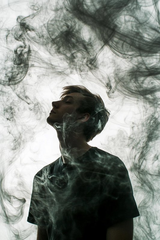 Silhouetted young man tilts head back eyes closed, surrounded by swirling wispy smoke against a b...