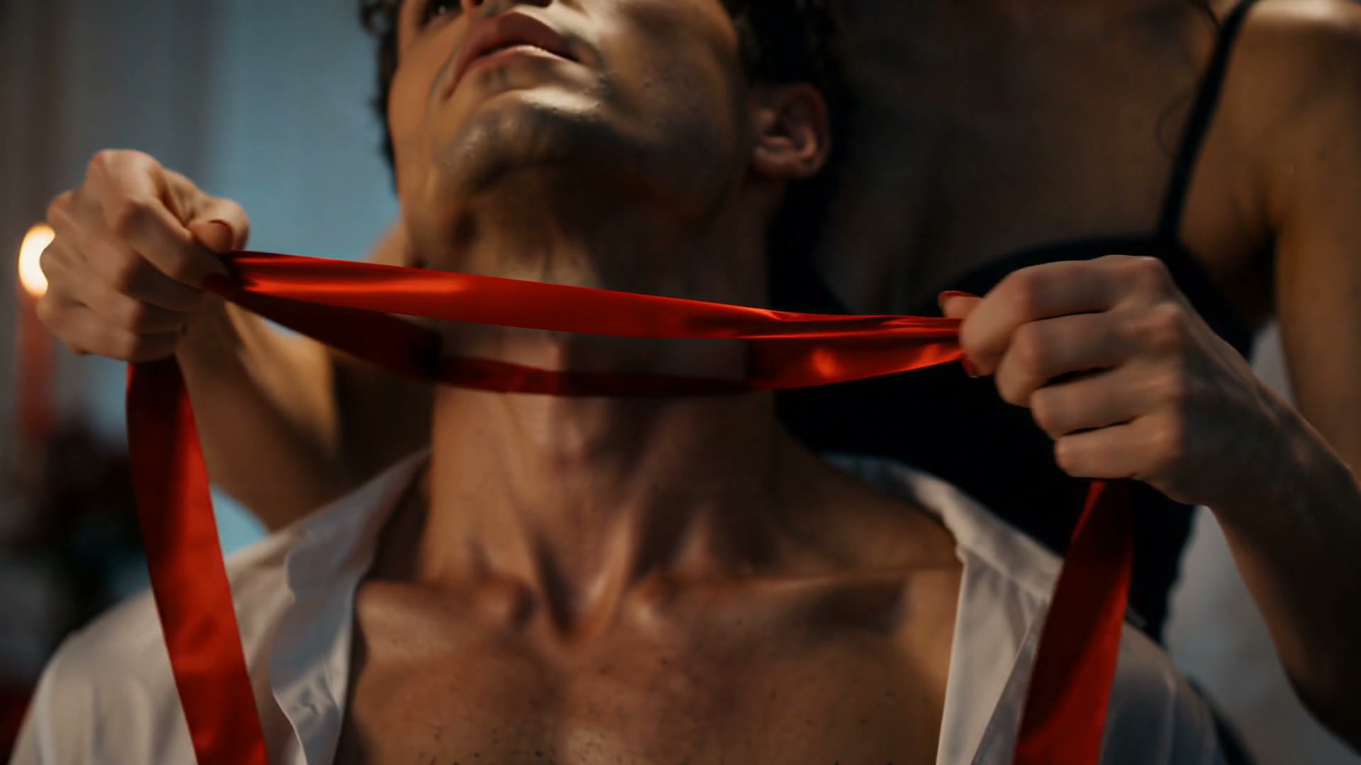 Woman holds glossy red satin ribbon around a man's neck in an intimate, moodily lit romantic closeup