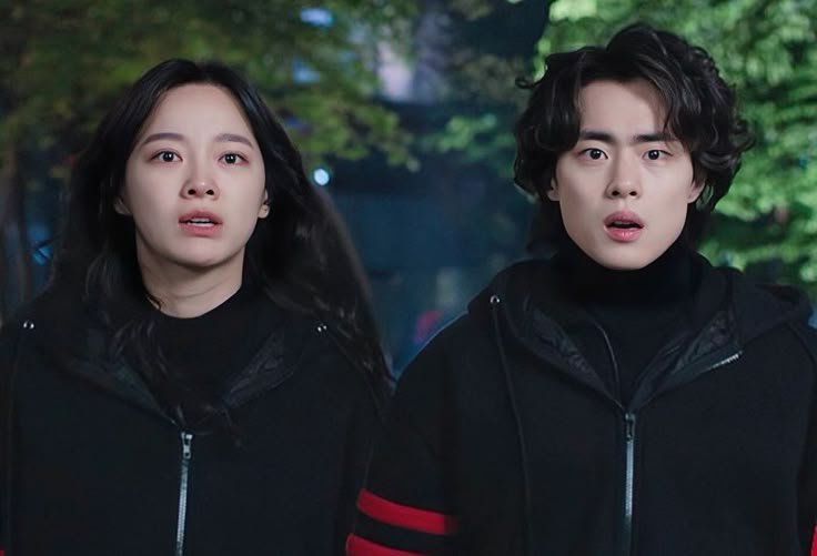 Two young people in black jackets with surprised expressions outdoors at night.
