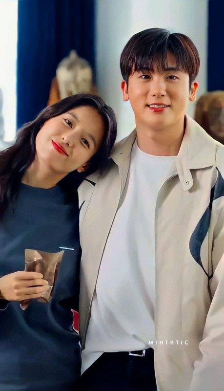 Two young adults smiling, woman leaning on the man’s shoulder in an indoor casual setting.