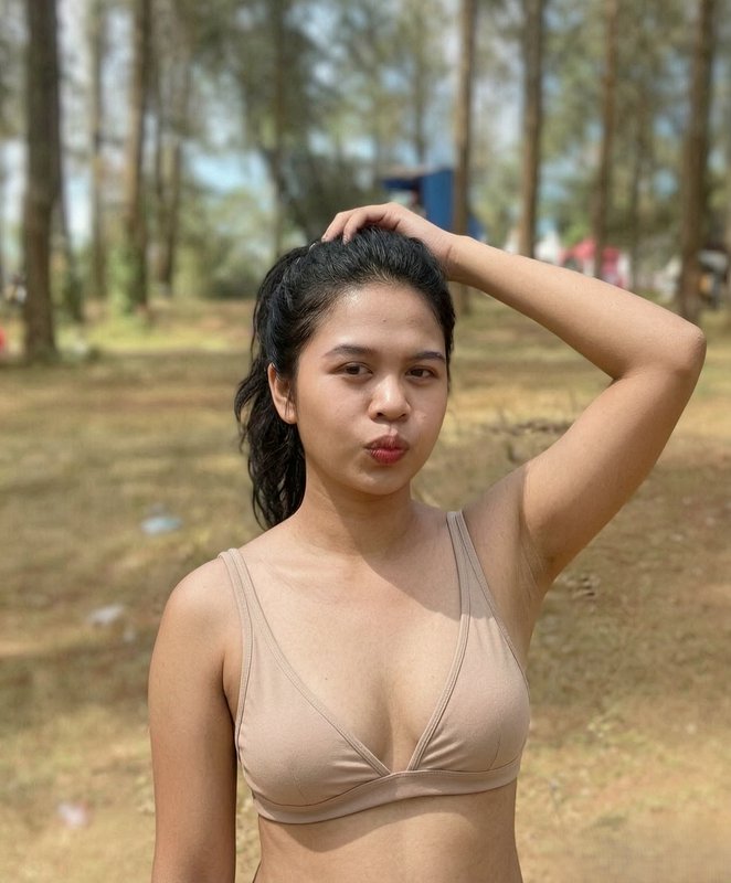 A young woman in a beige bikini top outdoors, lifting her hair with one hand.