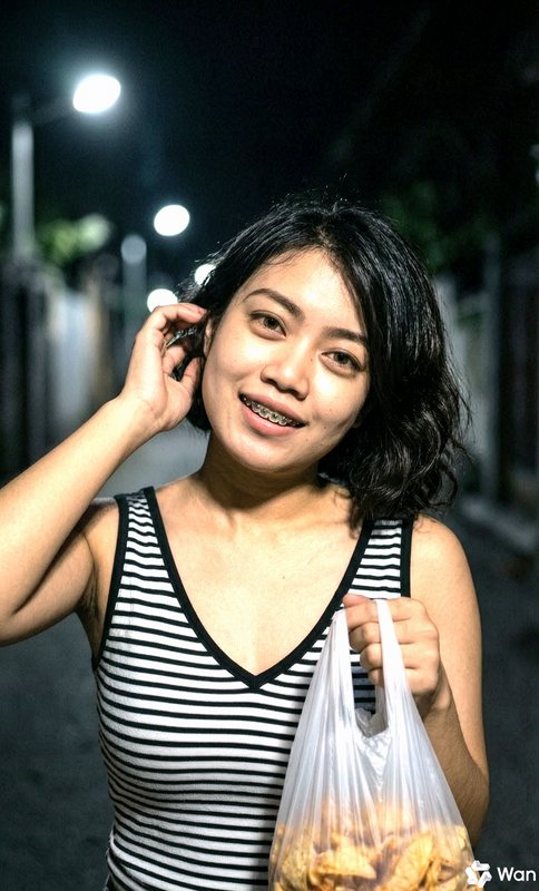Young woman with braces smiling at night, wearing a striped tank top and holding a plastic bag of...