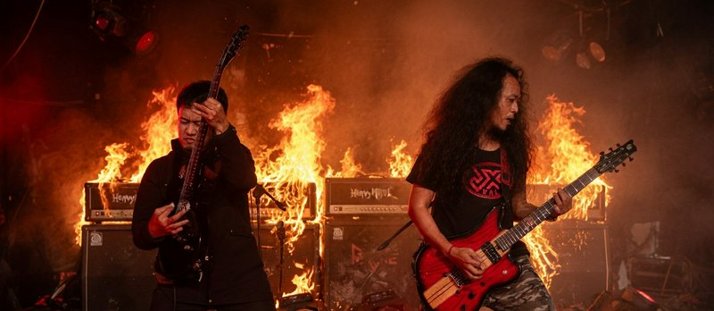 Two guitarists perform on stage with intense flames in the background during a metal concert.
