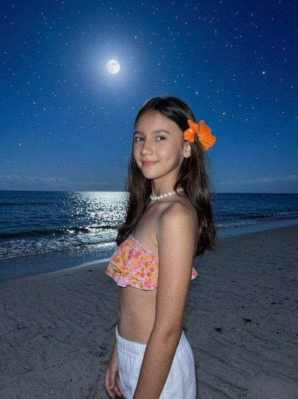 Young girl on a moonlit beach wearing a pink floral bikini top and an orange flower in her hair.