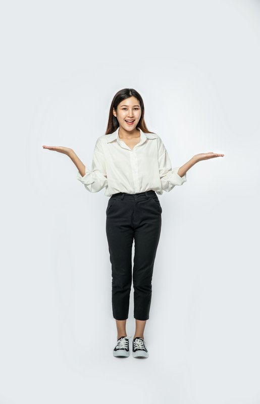 Young woman in a white shirt and black pants stands with arms outstretched against a white studio...