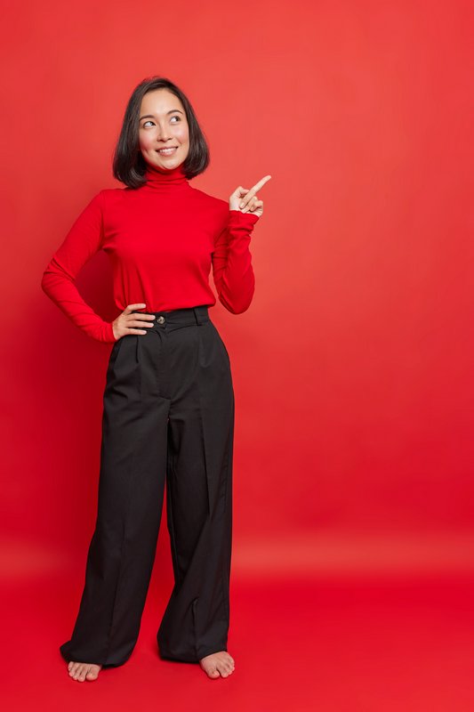 Woman in red turtleneck and black wide-leg pants posing against a red backdrop, barefoot.