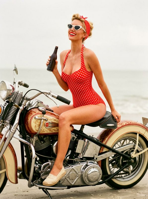 Pin-up style woman in a red polka-dot swimsuit on a vintage motorcycle at the beach, holding a bo...