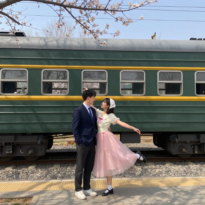 Couple in vintage outfits posing by a green train car with cherry blossoms overhead