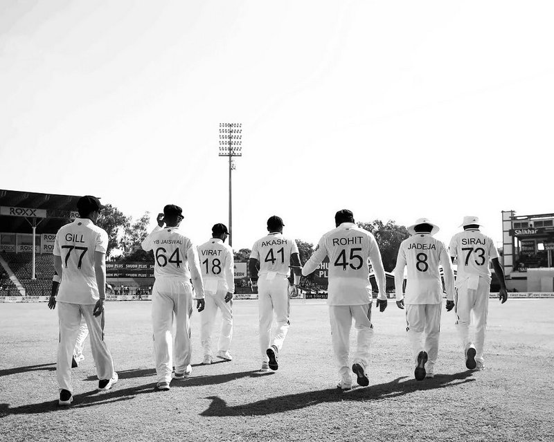 Cricket players in white uniforms walking on a field, backs with names and numbers visible.