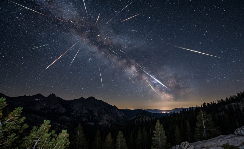 Night sky with Milky Way and meteors over a forested mountain landscape.