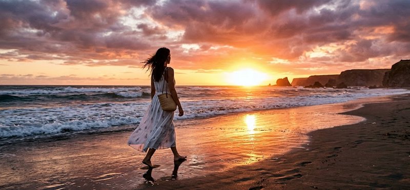 Woman in a flowy white dress walks barefoot along a dark sand ocean beach during a dramatic golde...