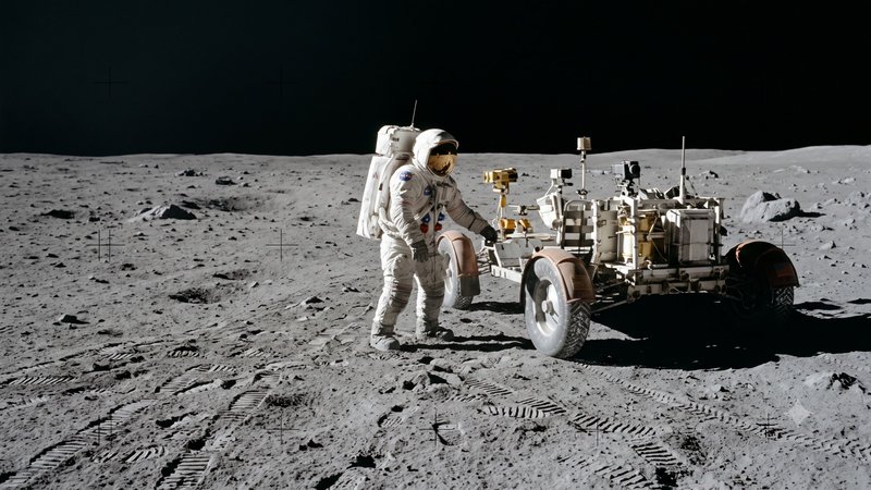 NASA astronaut in a white spacesuit stands next to an Apollo lunar rover on dusty, rocky moon ter...