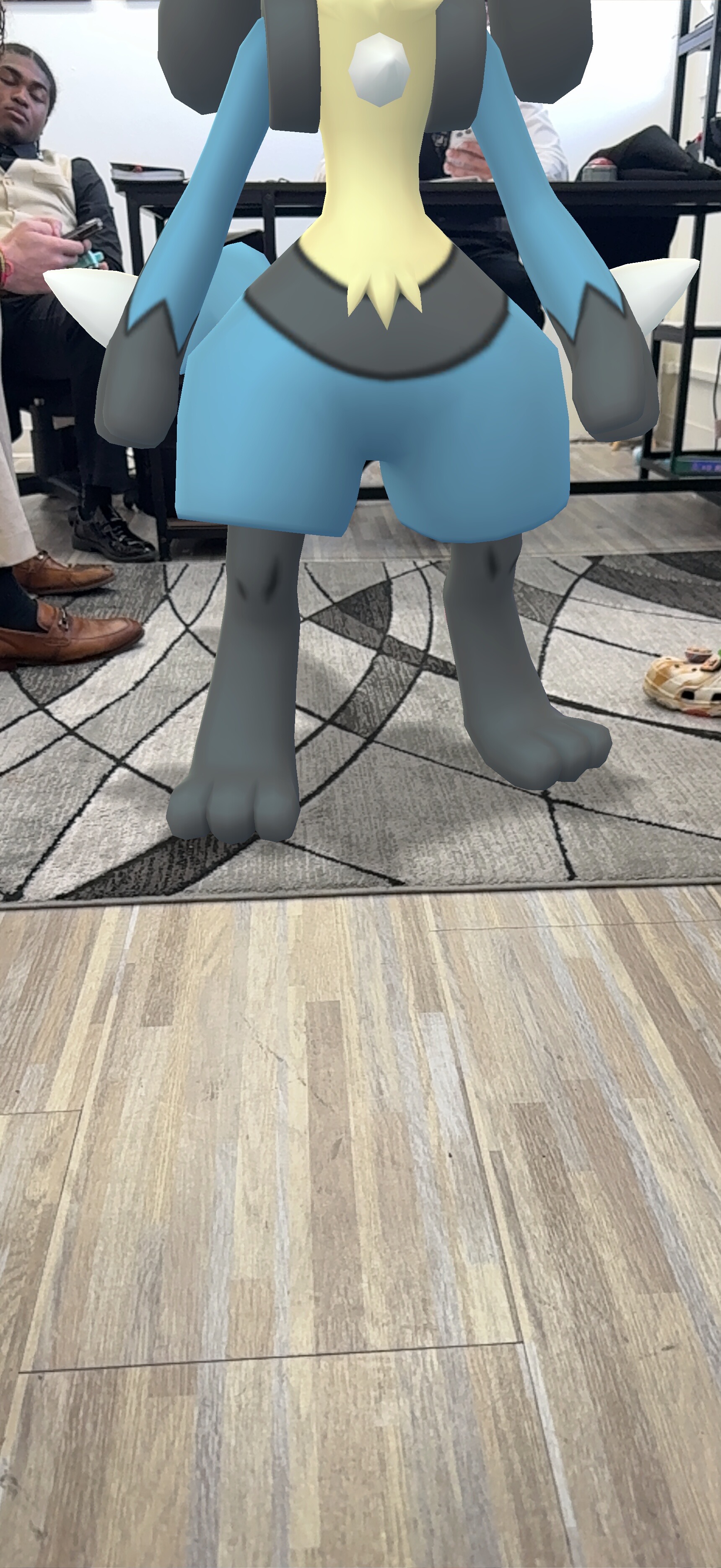 Augmented reality creature standing indoors on a patterned rug, blue and gray body with yellow chest.