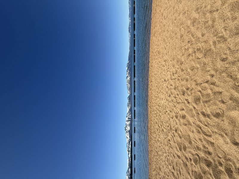Sunlit sandy beach with footprints along the shore, calm water, and snow-capped mountains in the distance under a clear