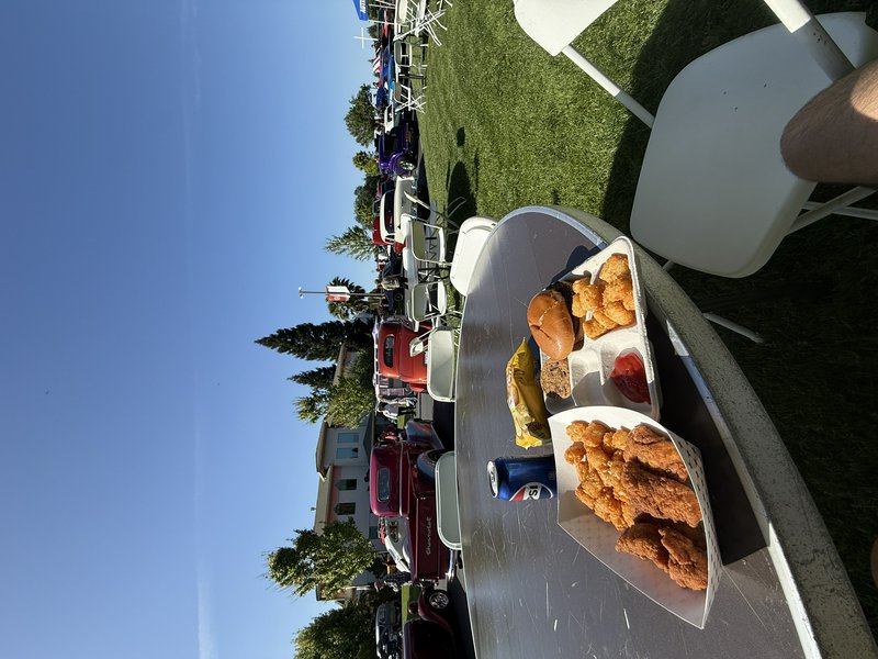 Outdoor picnic table with fried chicken, burgers, fries and sauces at a sunny car show on a grass...