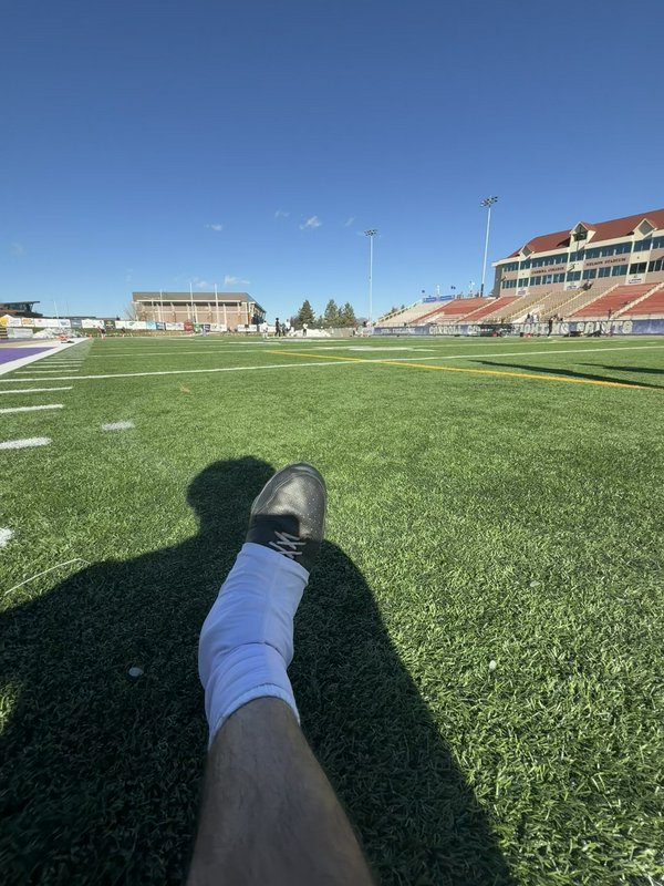 A football field view from the turf with a leg resting on the grass and bleachers in the background.