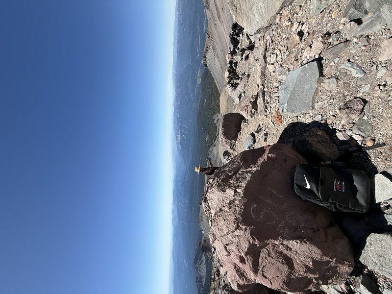 Rocky mountain summit with a backpack resting on a boulder overlooking a valley under a clear blue sky.