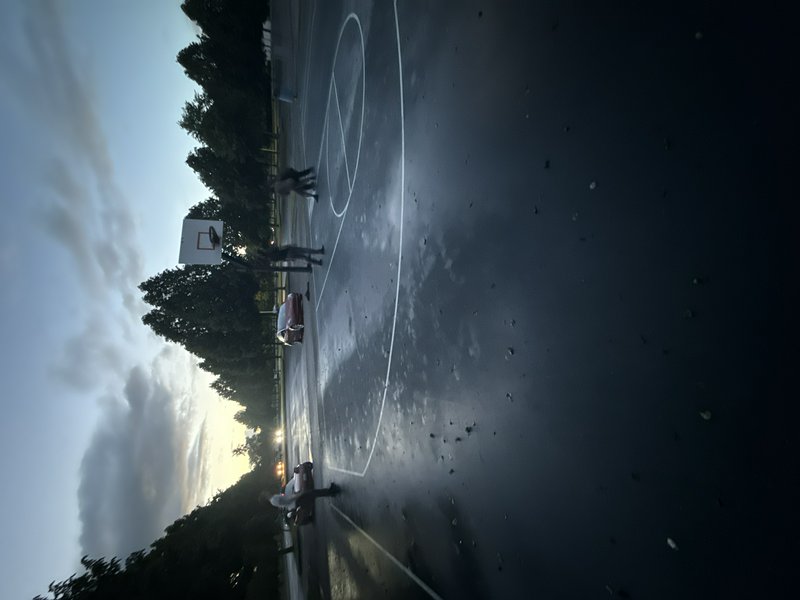 Outdoor basketball court after rain with players and a wet, reflective surface at dusk.