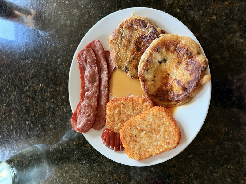 Overhead shot of a white breakfast plate with blueberry pancakes, bacon, hash browns, maple syrup...