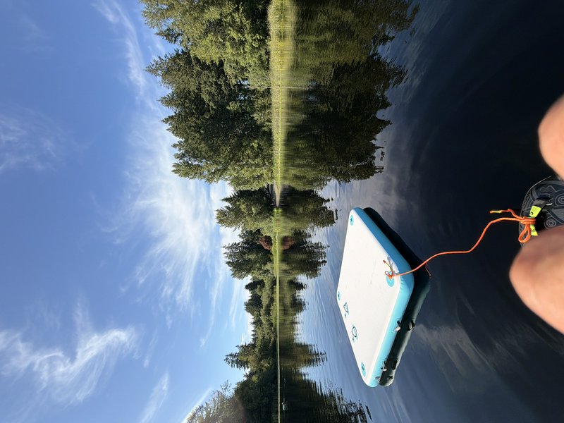 First person POV of calm forest lake, floating inflatable swim platform, tree reflections, bright...
