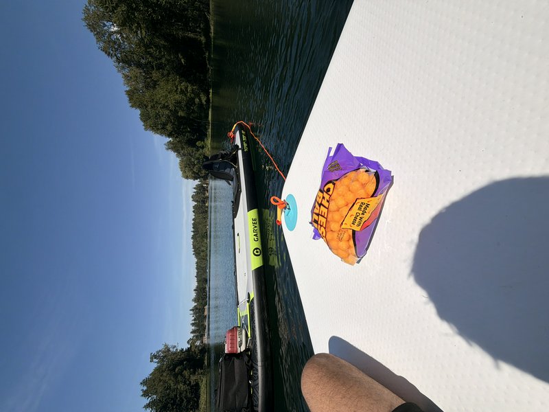 First person POV from a stand up paddleboard on a sunny lake, open cheese puff snack, tied inflat...