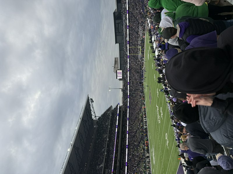 Elevated sideline view of a packed college football stadium, bundled fans overlooking green turf ...