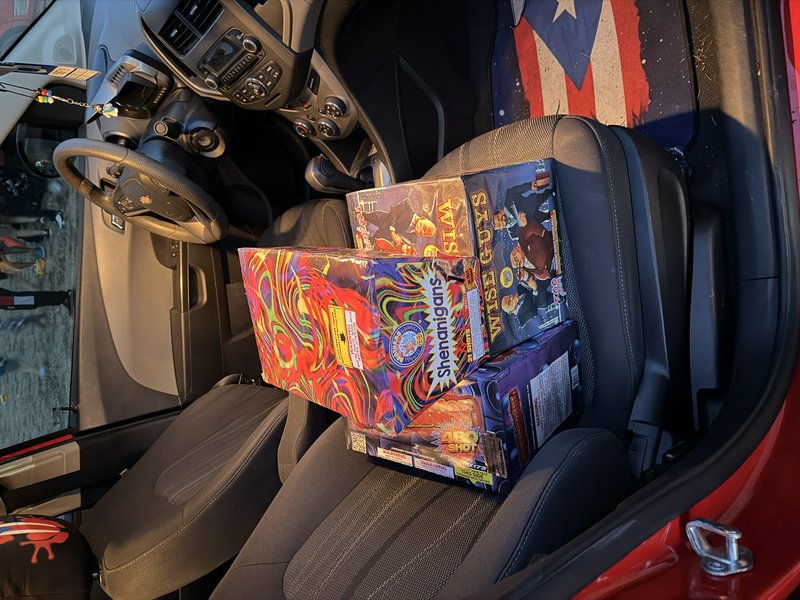 Stacked colorful fireworks boxes resting on the front passenger seat inside a compact car.