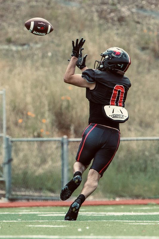 American football player #0 reaches up to catch an airborne pass on a green turf football field.