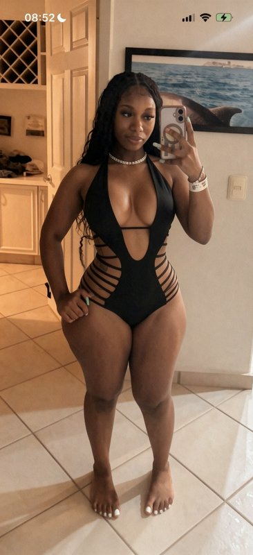 Black woman taking mirror selfie in a strappy cutout black halter one-piece swimsuit inside a sun...