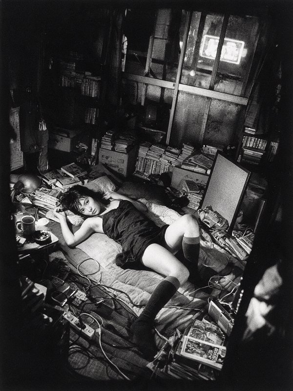 High-angle black and white shot of young woman looking at camera, reclining on cluttered bed in cramped Japanese apartment