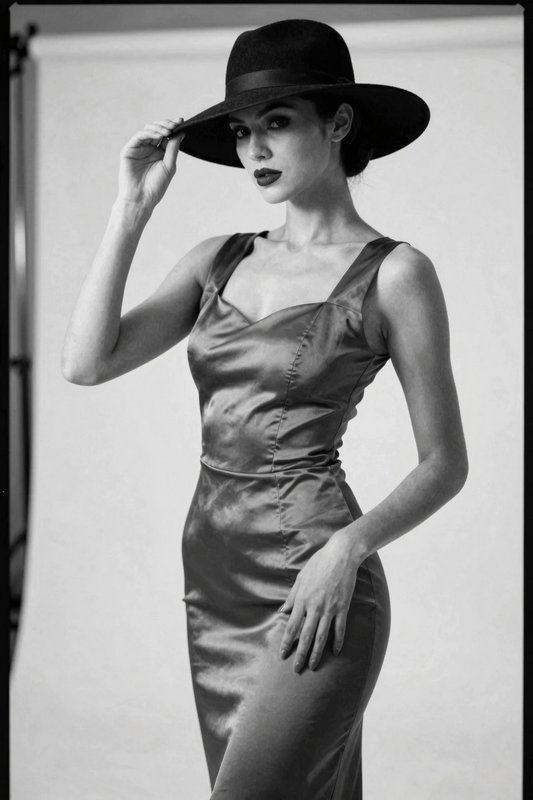 Elegant woman tipping her wide brim hat while wearing a formfitting satin dress in a studio portrait