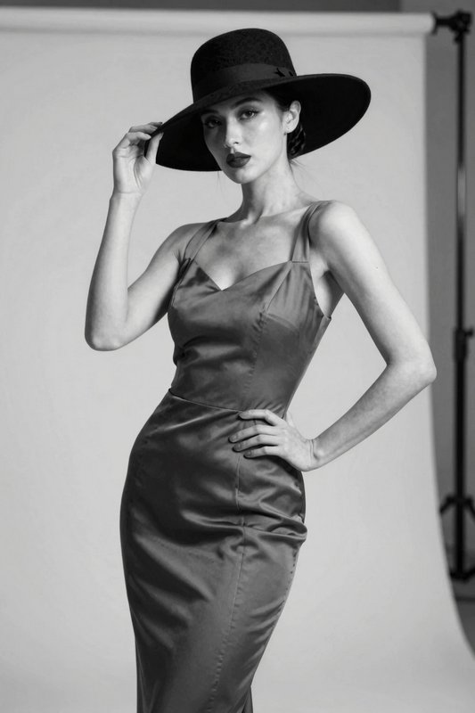 Elegant woman in a satin slip dress tipping her wide-brimmed hat in a monochrome studio portrait