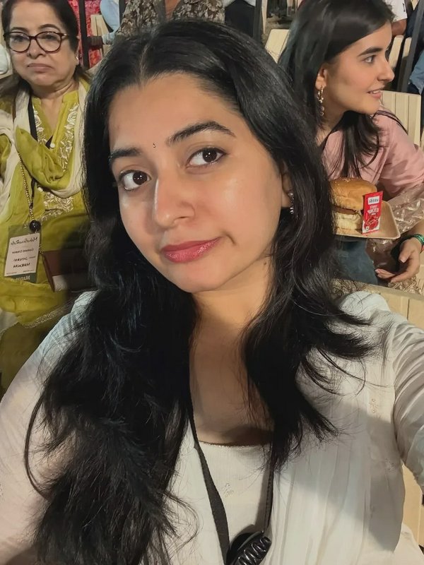 A young woman with long dark hair taking a selfie at an event, with others seated in the background.